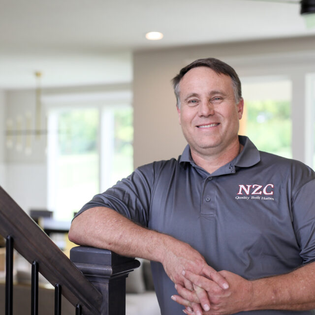 A man in a gray shirt standing in front of a staircase.